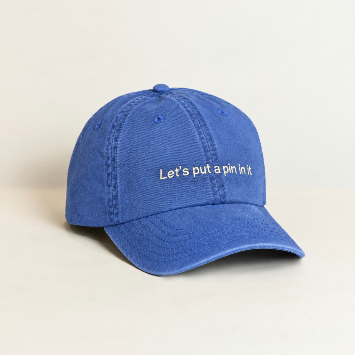 A blue cotton baseball cap with the phrase 'Let's put a pin in it' embroidered in white on the front, set against a plain beige background.