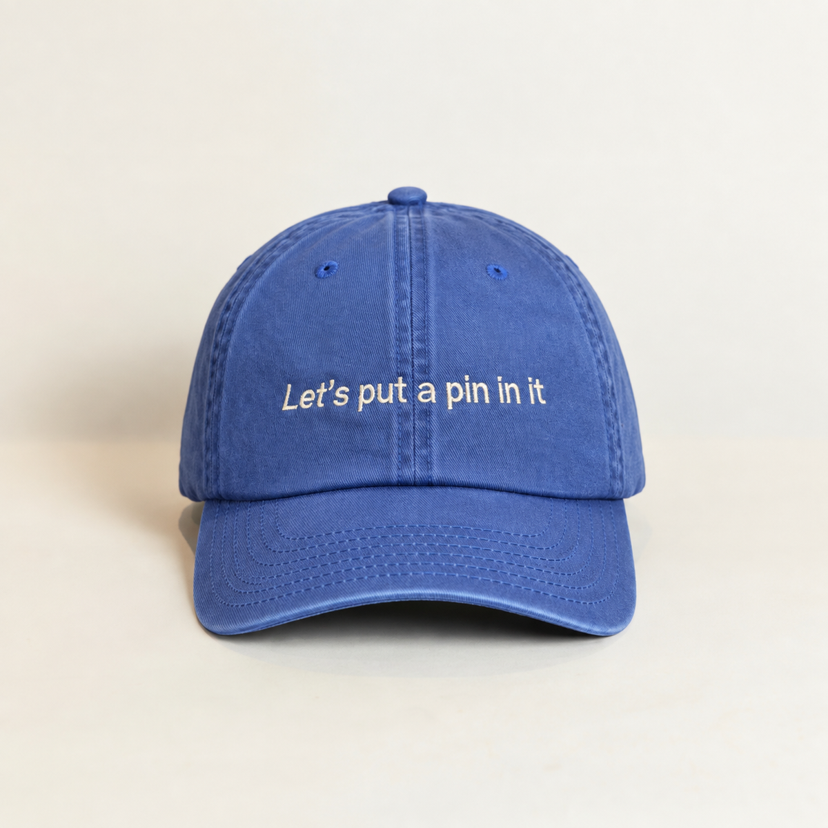 A blue cotton baseball cap with the phrase 'Let's put a pin in it' embroidered in white on the front, set against a plain light beige background.