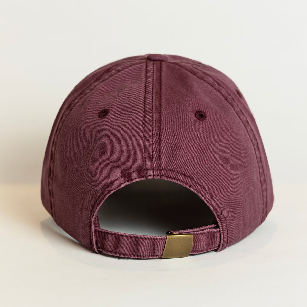 A washed mauve-purple baseball cap shown from the back, featuring a curved brim, ventilation eyelets, and an adjustable strap with a small gold-tone metal buckle.